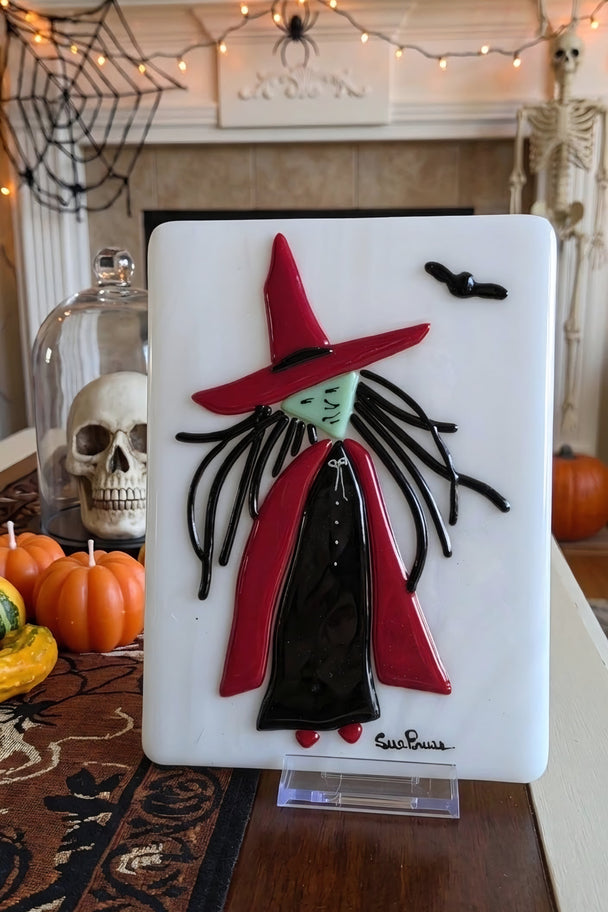 A Halloween-themed decorative wall plaque featuring a stylized witch with a red hat and cape on a white background, displayed on a tabletop in a decorated home setting with pumpkins, a skeleton, and a fireplace mantel.